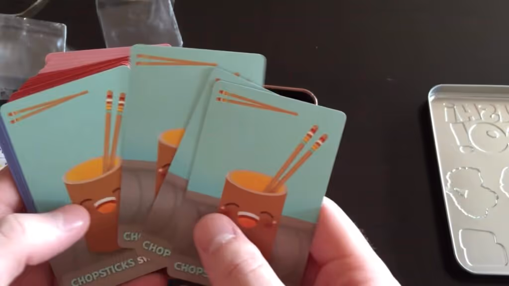 Hands holding Sushi Go! cards featuring chopsticks and a smiling cup